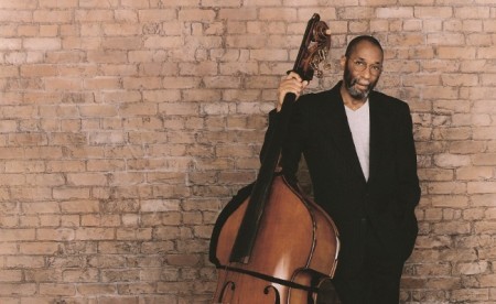 An image of Ron Carter standing by a brick wall with an upright bass.