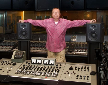 An image of Geoff Emerick in a red shirt in between two speakers and an old musical mixing console.