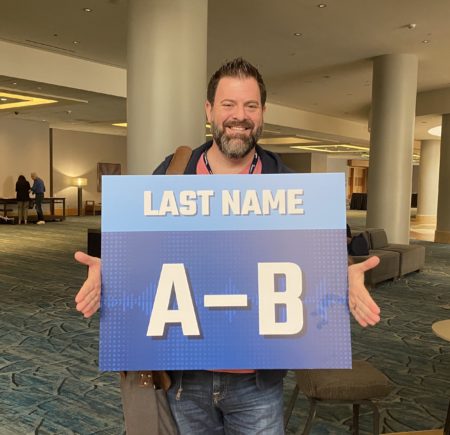 loren weisman holding a sign that reads last name, a to b with a hotel in the background.